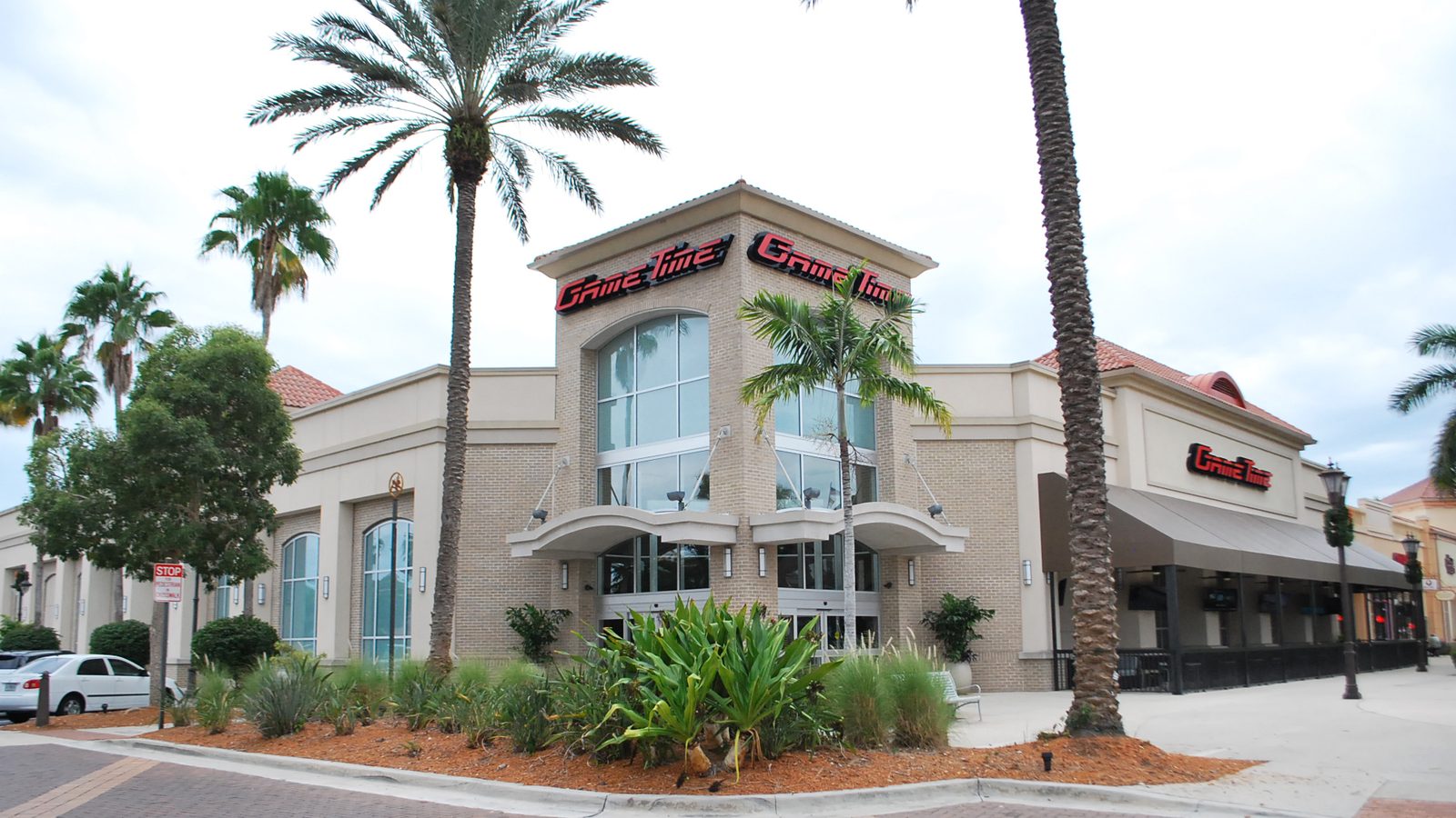 The outside of GameTime Fort Myers: a tan stucco building with a tall glass entrance, the red GameTime sign above it, palm trees and a planted bed in front