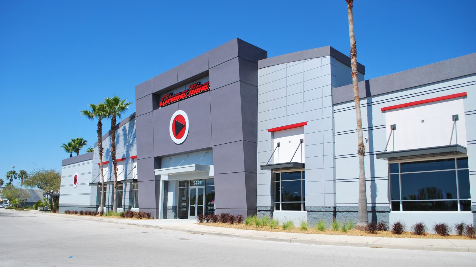 The outside of GameTime Ocoee from the street: a gray building with the red play-button logo above the glass entrance, red-trimmed windows and palm trees behind